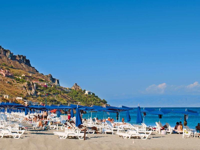 Strand met ligstoelen en blauwe parasols aan een kustlijn met heuvels onder een heldere hemel