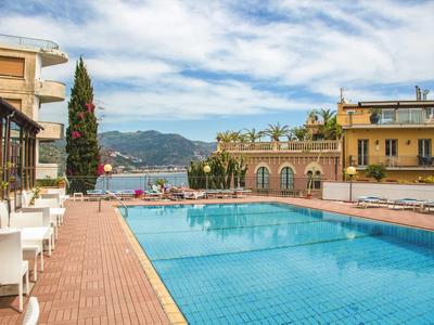 Ein klarer blauer Pool mit Liegestühlen, umgeben von Gebäuden und Bergen im Hintergrund.