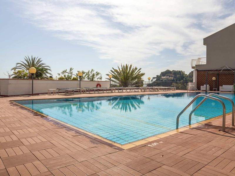 Buitenzwembad op terras met zonnescherm en palmbomen op de achtergrond onder een heldere lucht.