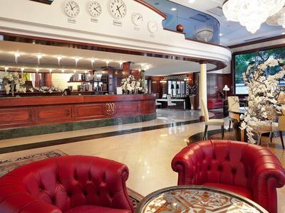 Elegancki hol hotelowy z czerwonymi skórzanymi fotelami, recepcją i dekoracyjnym dywanem.