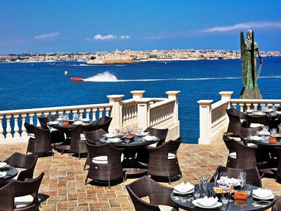 Taras z stolikami i krzesłami z widokiem na morze i odległe miasto.