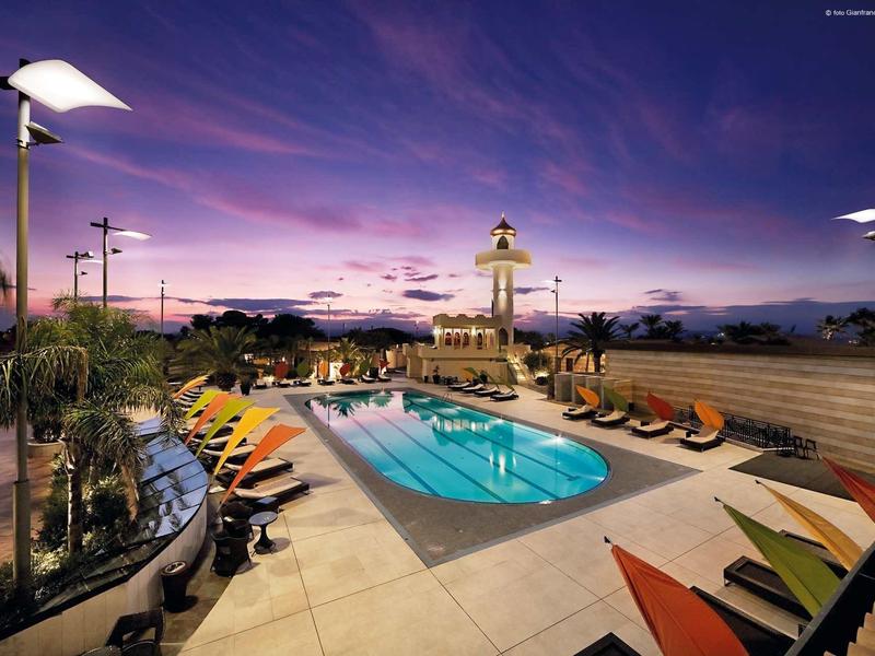 Wieczorny widok oświetlonego basenu z kolorowymi leżakami i palmami pod dramatycznym niebem.