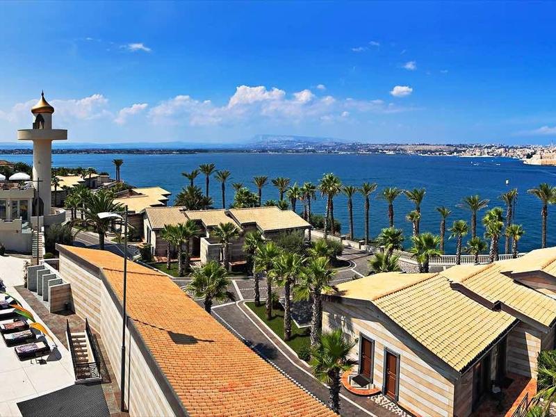 Widok na basen hotelowy i palmy nad morzem pod jasnym niebieskim niebem z portem w tle.