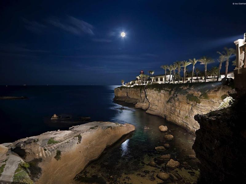 Nadmorskie klify nocą z blaskiem księżyca i podświetlonymi budynkami w tle.