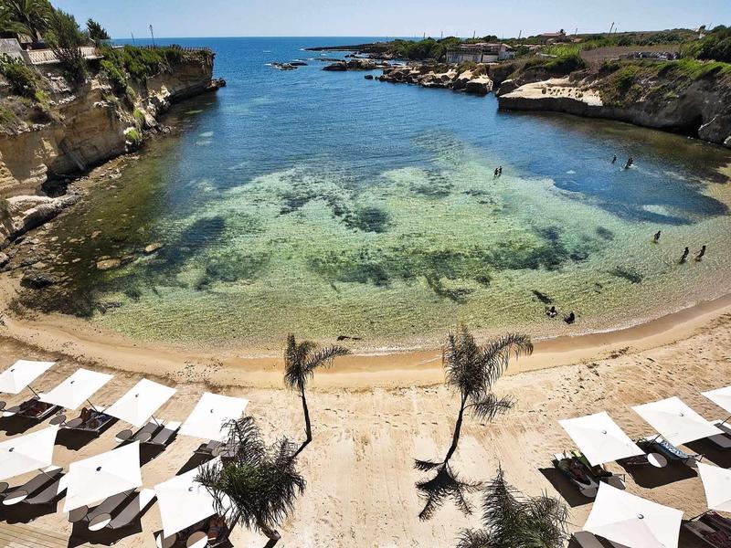 Widok na spokojną zatokę z czystą wodą, piaszczystą plażą i parasolami przy basenie hotelowym.