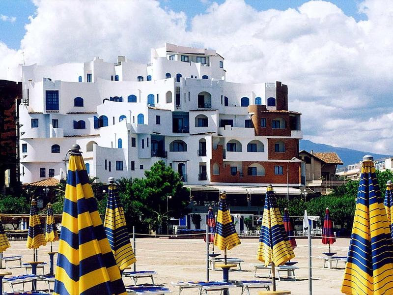 Spiaggia con ombrelloni gialli e blu davanti a un hotel bianco su più piani sotto un cielo nuvoloso.