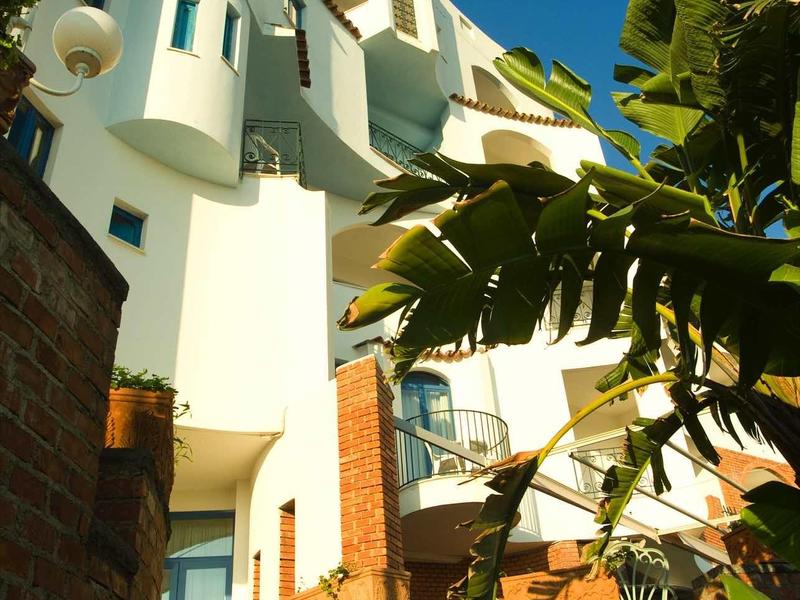 Edificio moderno di un hotel con pareti bianche, finestre tonde e piante tropicali in primo piano.