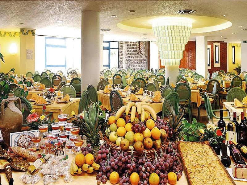 Ristorante luminoso e spazioso con buffet, esposizione di frutta e sedute con sedie verdi.