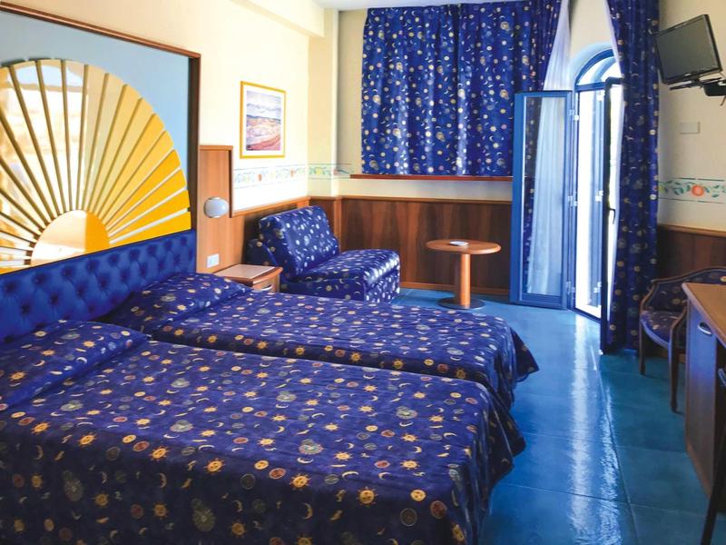 Camera d'hotel con due letti singoli, tende blu e zona salotto vicino alla finestra.