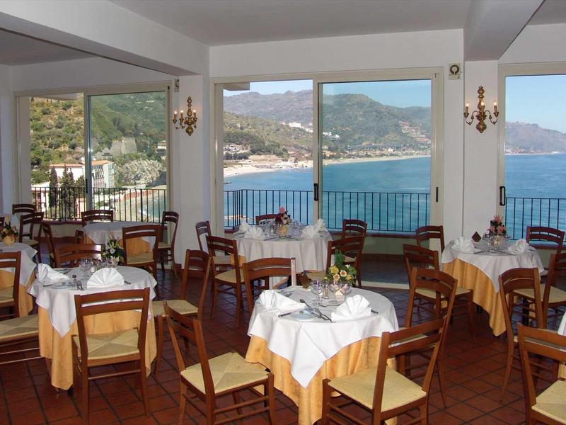 Restaurant with wooden tables and chairs, sea and mountain views through large windows.