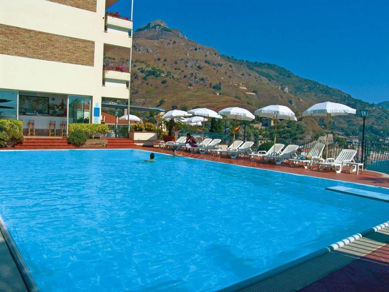 Hotel pool with loungers and umbrellas against mountainous background under clear blue sky.