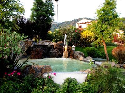 Una piccola piscina con cascata in un giardino rigoglioso con colline e case sullo sfondo.