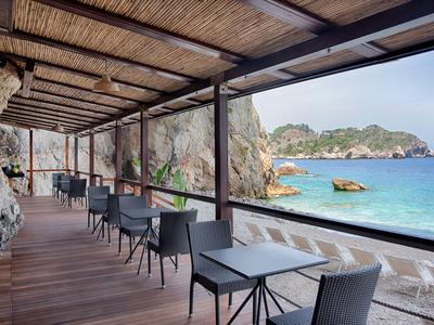 Terrazza aperta con tavoli e sedie sul mare, circondata da rocce e acqua limpida.