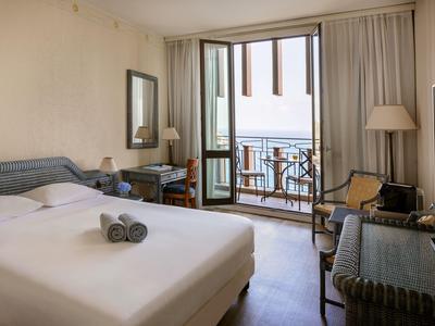 Camera d'hotel luminosa con letto matrimoniale, scrivania e balcone con vista mare.