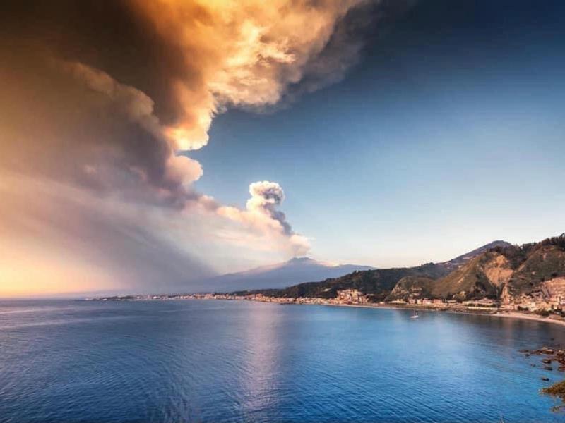 Landschap met vulkaan die rookwolken boven de zee uitstoot bij zonsondergang.