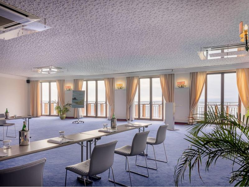 Hel verlichte vergaderruimte met stoelen, tafels en grote ramen met gordijnen.