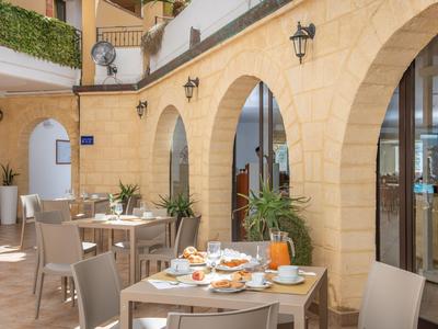 Sonnige Hotelterrasse mit Tischen, Stühlen und Frühstücksgedecken unter steinernen Bögen.