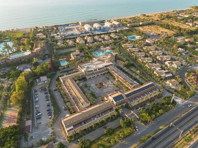 Luftaufnahme eines großen Hotelkomplexes mit Pool, umgeben von Grünflächen und Straße am Meer.