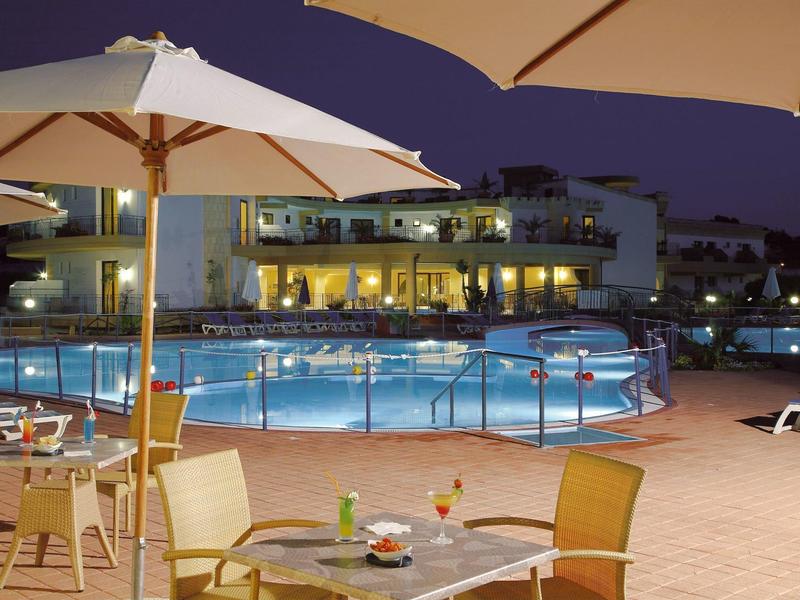 Evening pool area with illuminated lounge chairs and tables under umbrellas at a hotel.