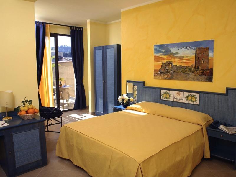 Bright hotel room with double bed, yellow walls, blue furniture, and balcony with view.