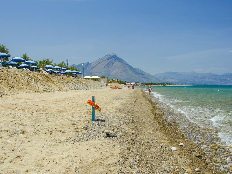 Beach with lounge chairs and umbrellas, mountain in the background, and blue sea under clear sky.