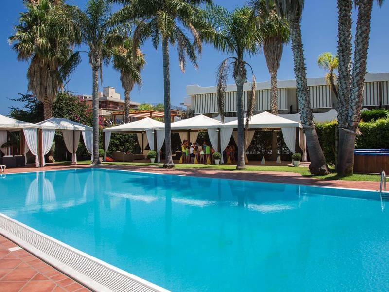 Großer blauer Pool mit weißen Sonnenschirmen und Palmen bei strahlendem Sonnenschein.