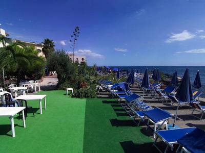 Strand mit blauen Liegestühlen und weißen Tischen neben einem Hotel unter blauem Himmel.