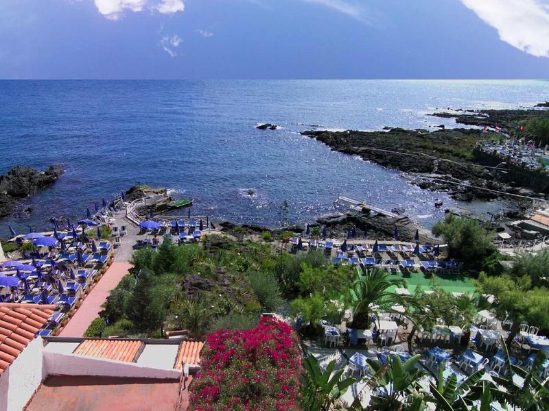 Vista di una zona costiera con hotel, piscina, lettini e vegetazione fiorita vicino al mare.