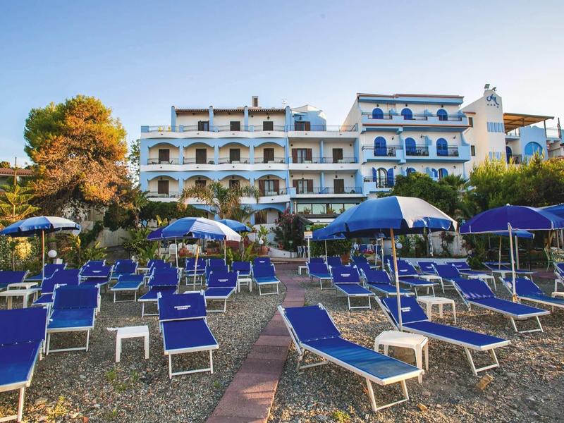 Spiaggia con lettini blu e ombrelloni davanti a un edificio dell'hotel bianco con accenti blu.