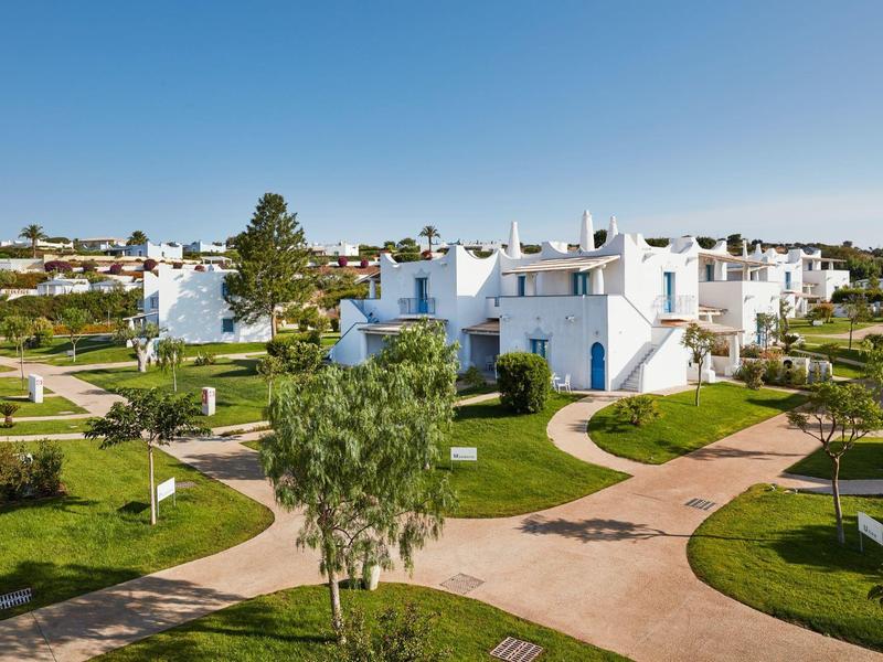 Maisons blanches dans un jardin verdoyant sous un ciel bleu, des chemins les relient.