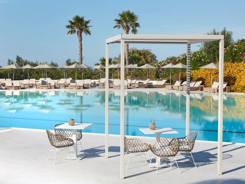 Espace piscine moderne avec fauteuils suspendus, palmiers et nombreuses chaises longues sous des parasols blancs.