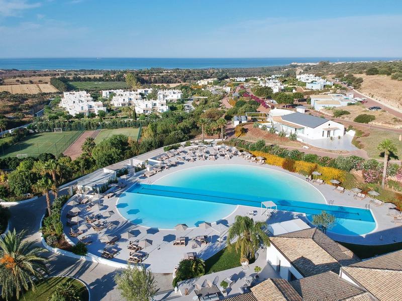 Grande piscine d'hôtel avec transats et parasols, entourée de bâtiments et vue sur la mer.