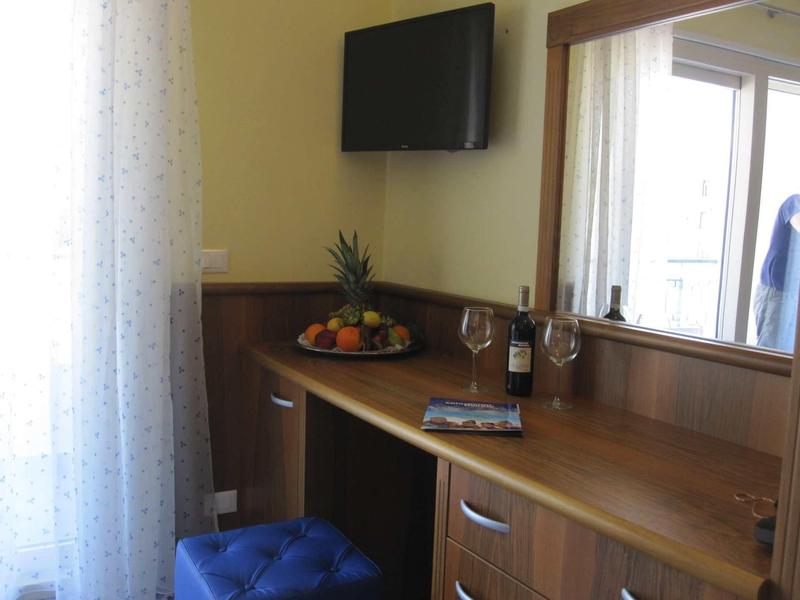 Chambre d'hôtel lumineuse avec bureau en bois, TV, corbeille de fruits et vin sur la table.