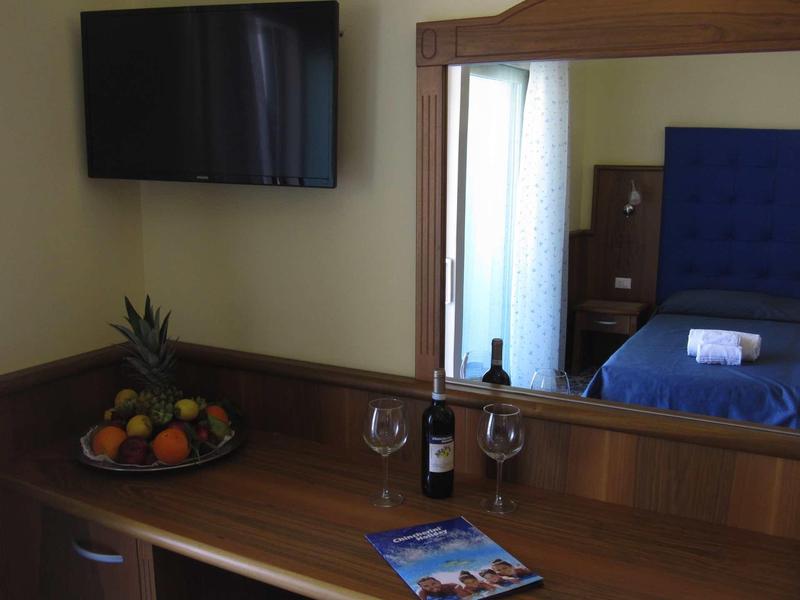 Chambre d'hôtel avec télévision, miroir, lit, bouteille de vin, verres à vin et plateau de fruits.