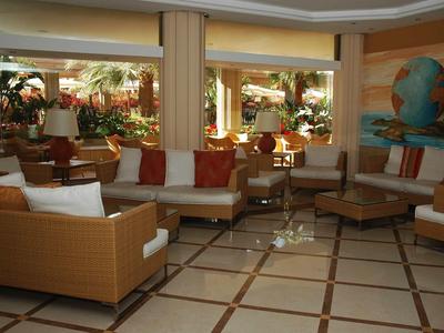 Salon d'hôtel lumineux avec canapés modernes, coussins et grandes fenêtres donnant sur des plantes.