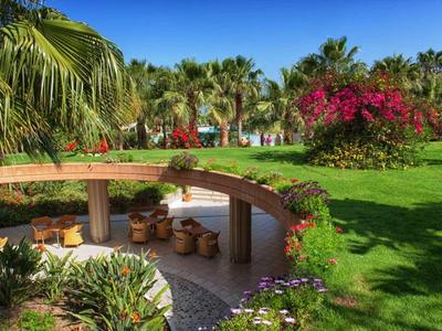 Jardin vert avec des fleurs, des palmiers et un coin salon avec table ronde en bois sous un ciel bleu