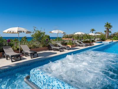 Blauer Whirlpool am Pool mit Liegestühlen und Sonnenschirmen vor Meerblick an sunnigem Tag.