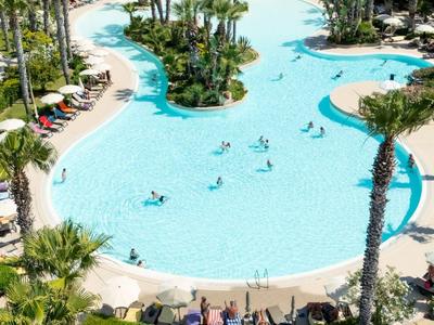 Großer Pool mit Palmen und Liegestühlen in einer Hotelanlage umgeben von Hügeln.