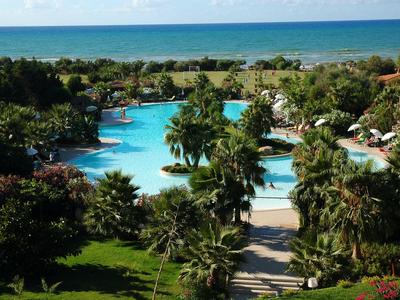 Großer Hotelpool mit umgebender Palmenlandschaft und Blick auf das blaue Meer.