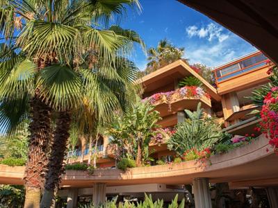 Hotel mit üppiger Vegetation, Palmen und blühenden Pflanzen unter blauem Himmel.