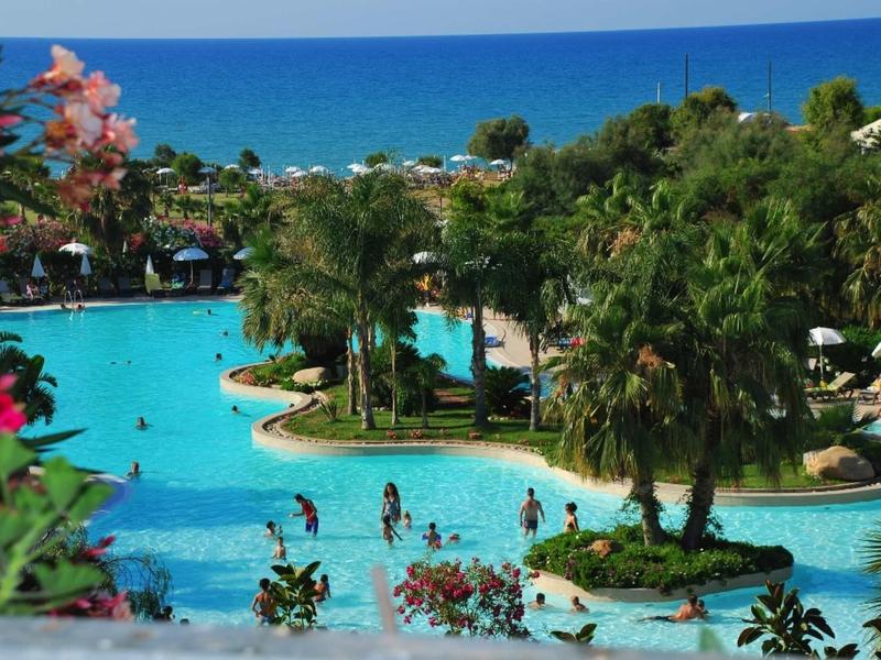 Grand piscine d'hôtel avec des palmiers et des invités, vue sur la mer bleue en arrière-plan.