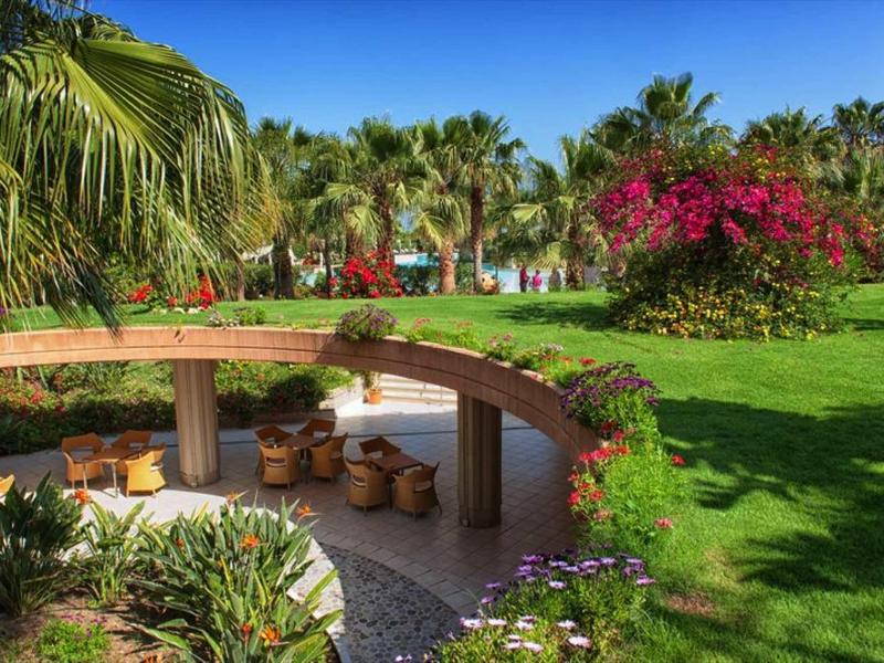 Jardin vert avec des fleurs, des palmiers et un coin salon avec table ronde en bois sous un ciel bleu