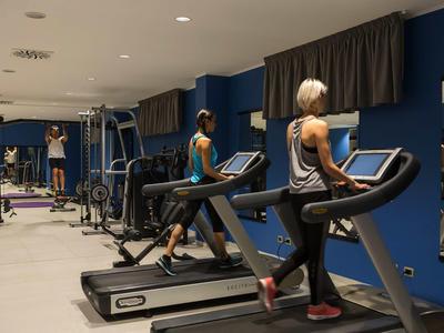 Dos mujeres hacen ejercicio en cintas de correr en un gimnasio moderno con paredes azules.