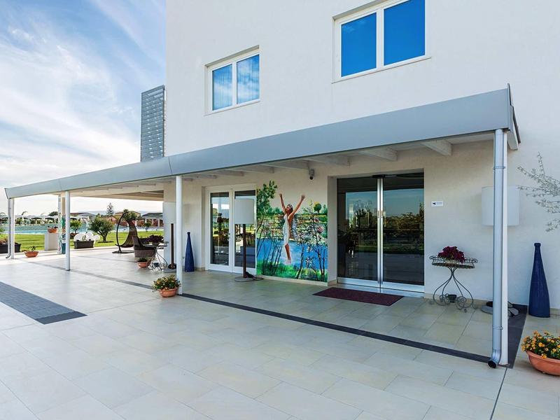 Moderní vstup do hotelu s krytou terasou a květináči pod jasnou oblohou.