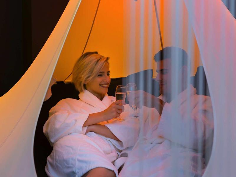 Žena v županu relaxuje s drinkem v útulném, barevně osvětleném prostředí.