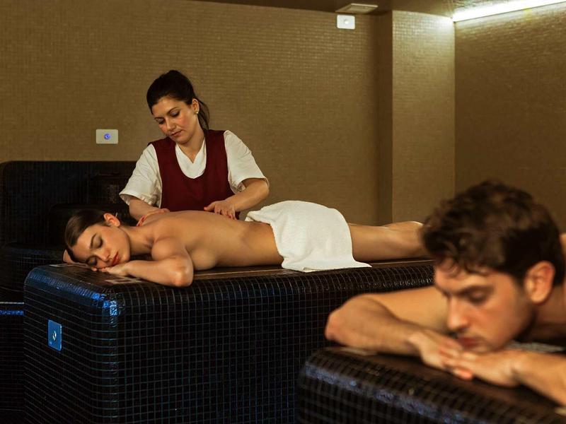 Dva lidé leží na masážních stolech, masérka provádí masáž zad v klidné místnosti.