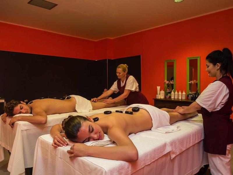 Dvě ženy dostávají masáže horkými kameny na bílých masážních stolech ve wellness místnosti.