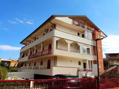 Mehrstöckiges Wohnhaus mit Balkonen, cremeweißer Fassade und rotem Zaun bei blauem Himmel.