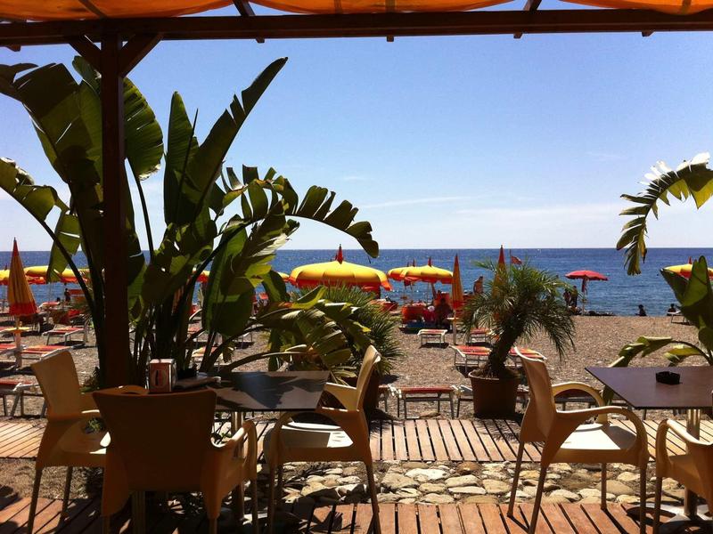 View from a shaded terrace with chairs and tables to a sunny beach with umbrellas and sea.