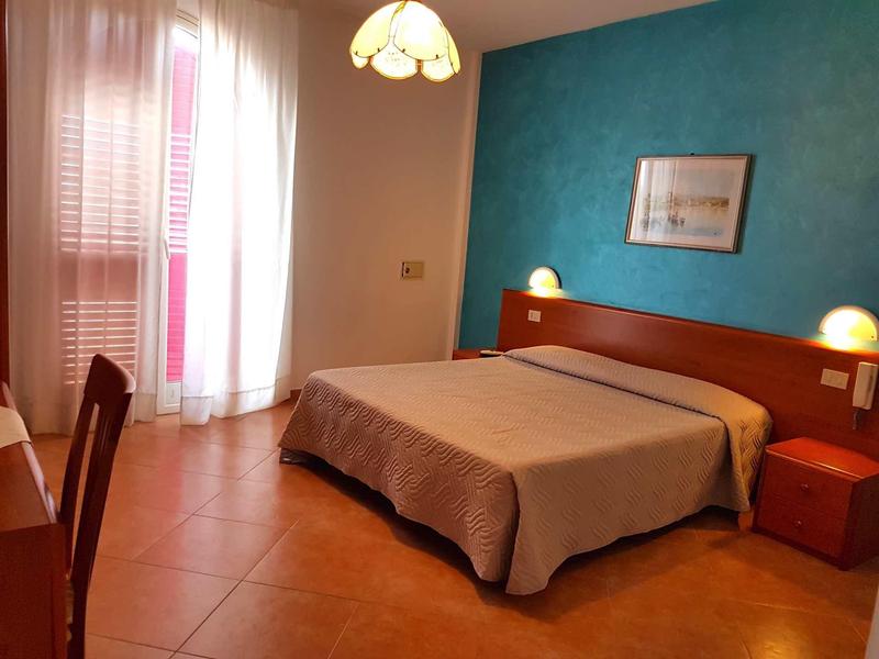 Brightly furnished hotel room with double bed, desk, chair, and daylight through window with curtains.